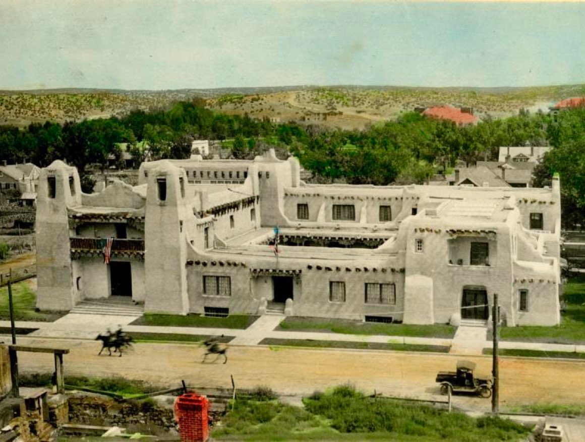 A high angle view of the museum with grey walls and a dirt road in front that has a car and horse riders on it