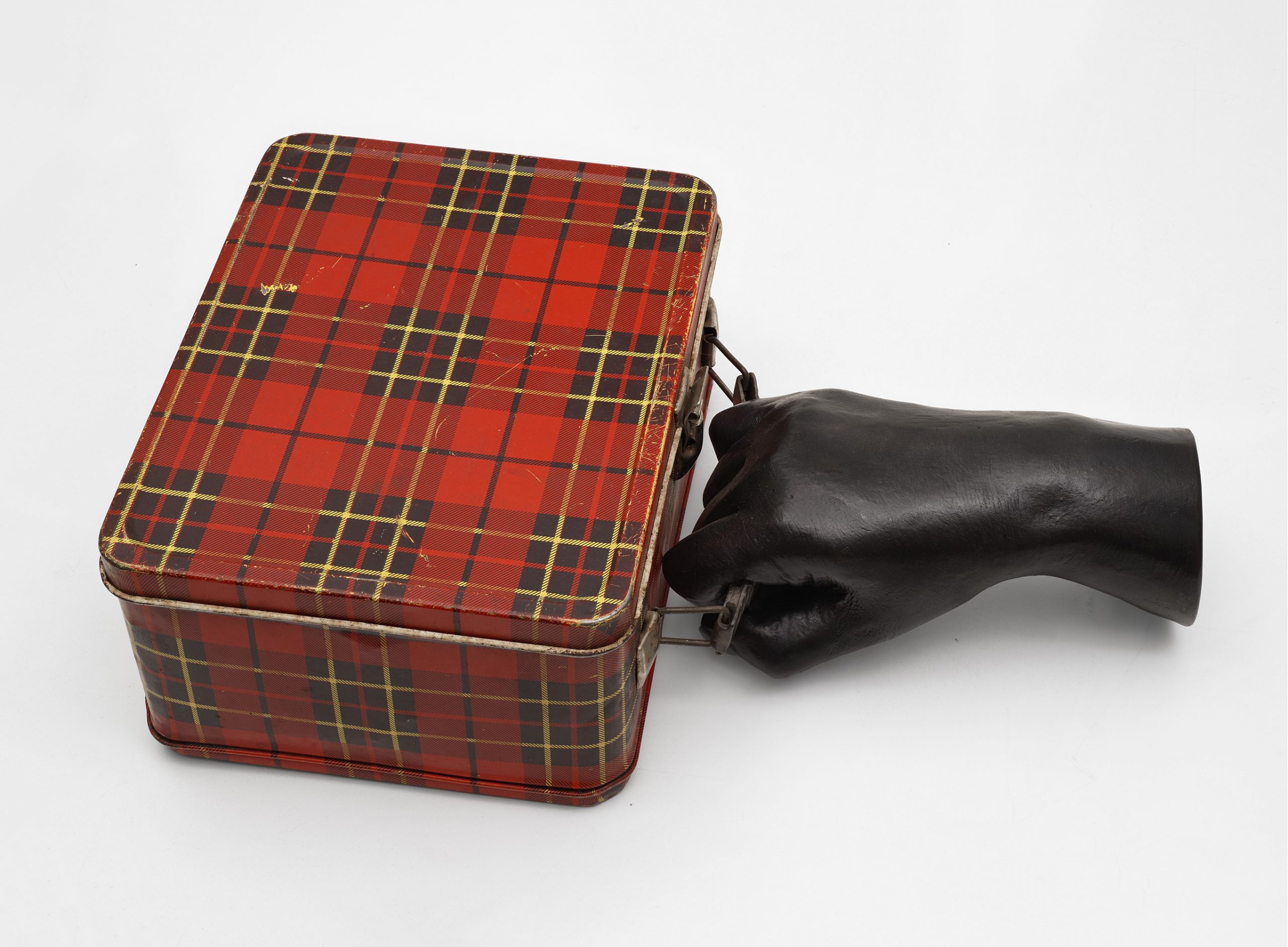 image of cast bronze hand in black, ending just above the wrist, gripping handle of square metal lunch box with red and green plaid design