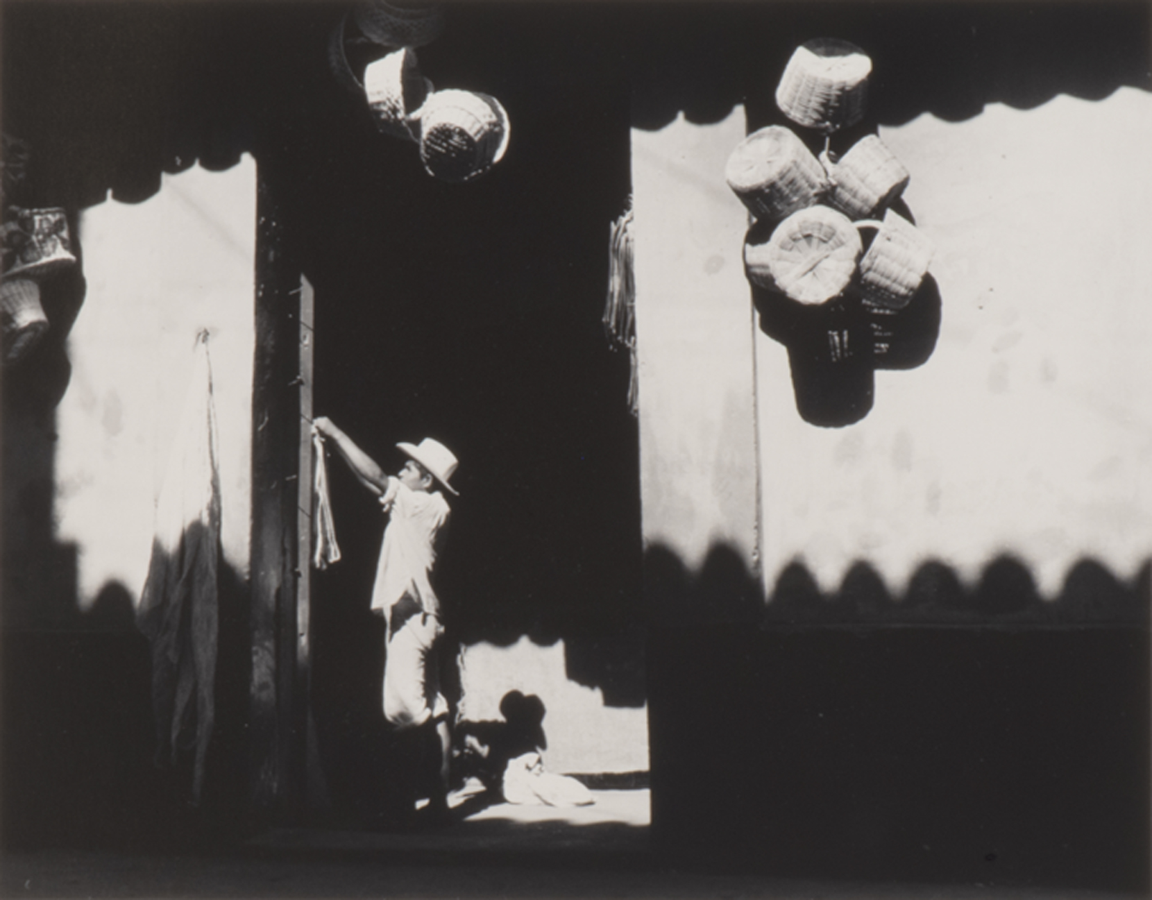 Man by doorway with baskets hanging on the outside wall, dark shadows of fence and roof line across bottom and top of photograph.