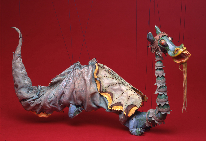 Marionette of large green dragon, with soft leather body and long jointed neck, large protruding jaw with dangling beard, long tail.