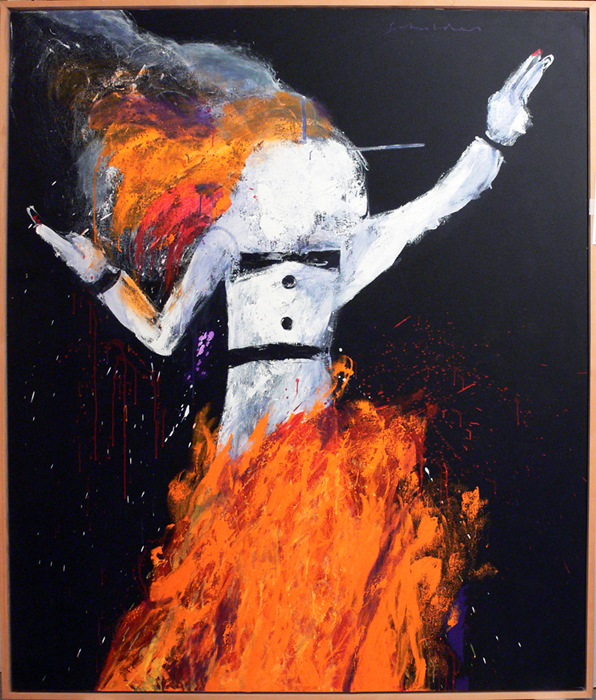 A white painted puppet of a human figure wearing a black bow tie and black belt (Zozobra (Old Man Gloom)) is on fire. Flames and smoke burst from the back of the puppet's head. The bottom half of the figure is burning with large orange flames reaching up to its waist. The background is black with specks of smoke and fire debris floating around the puppet.