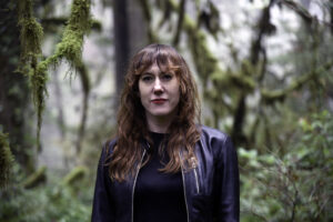 Portrait of a woman shown from the waist, wearing black shirt and black jacket with blurred green foliage in the background.