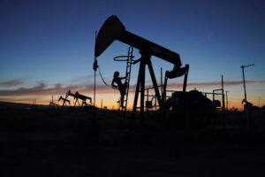 An oil derrick and pump is silhouetted against an orange and pink sunset. The silhouette of a woman with fringed sleeves is seen leaning halfway up the derrick's ladder in a sultry pose.