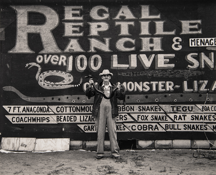 A man in a hat holding a snake stands in front of a large sign advertising the Regal Reptile Ranch