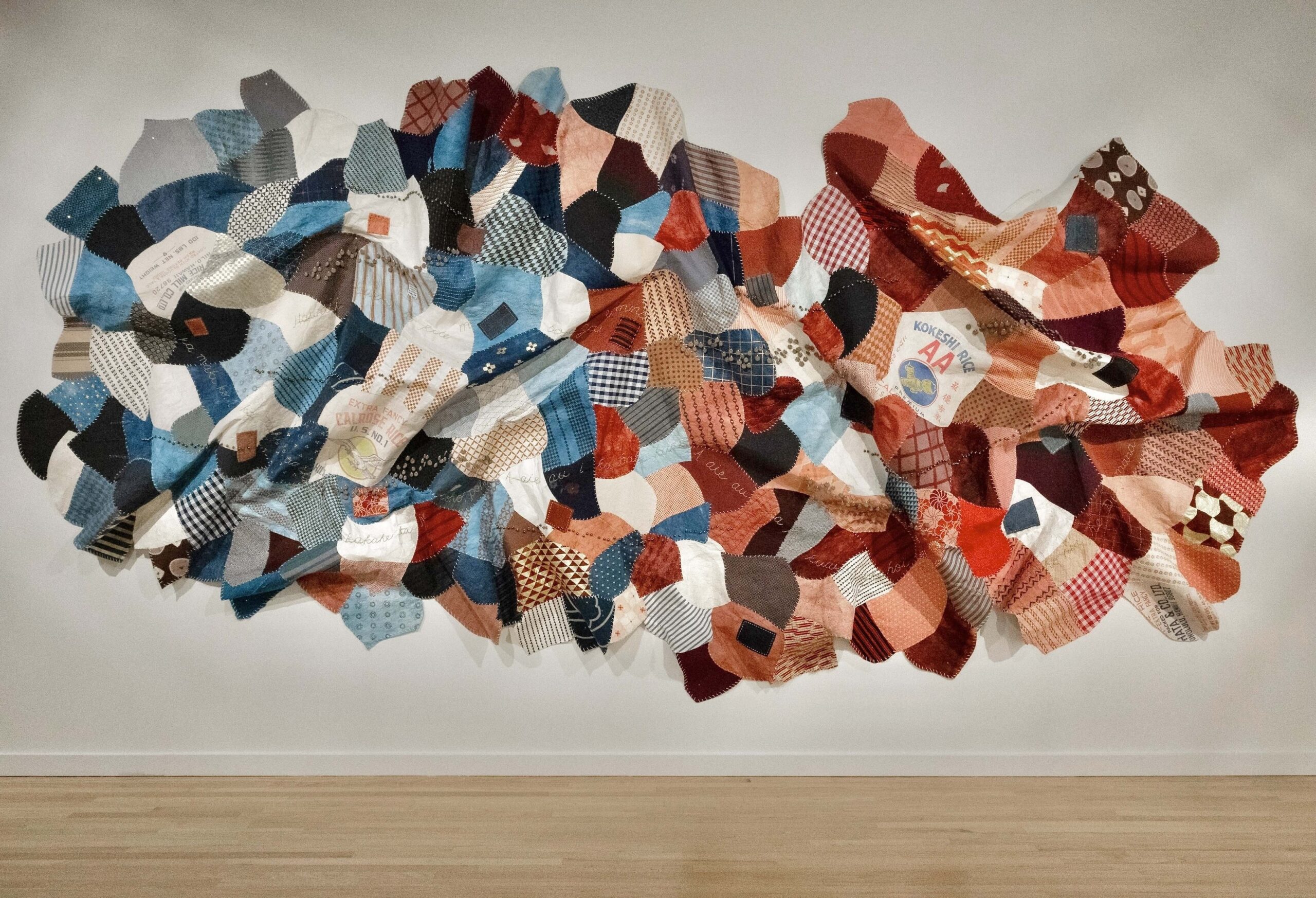A large, predominantly red and blue collaged textile artwork hangs on a wall