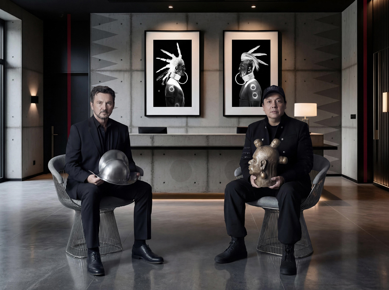 Two men sit in chairs in front of a wall with artwork on it. The man on the left holds a conquistador's helmet in his lap, and the man on the right holds a sculpture of a head.